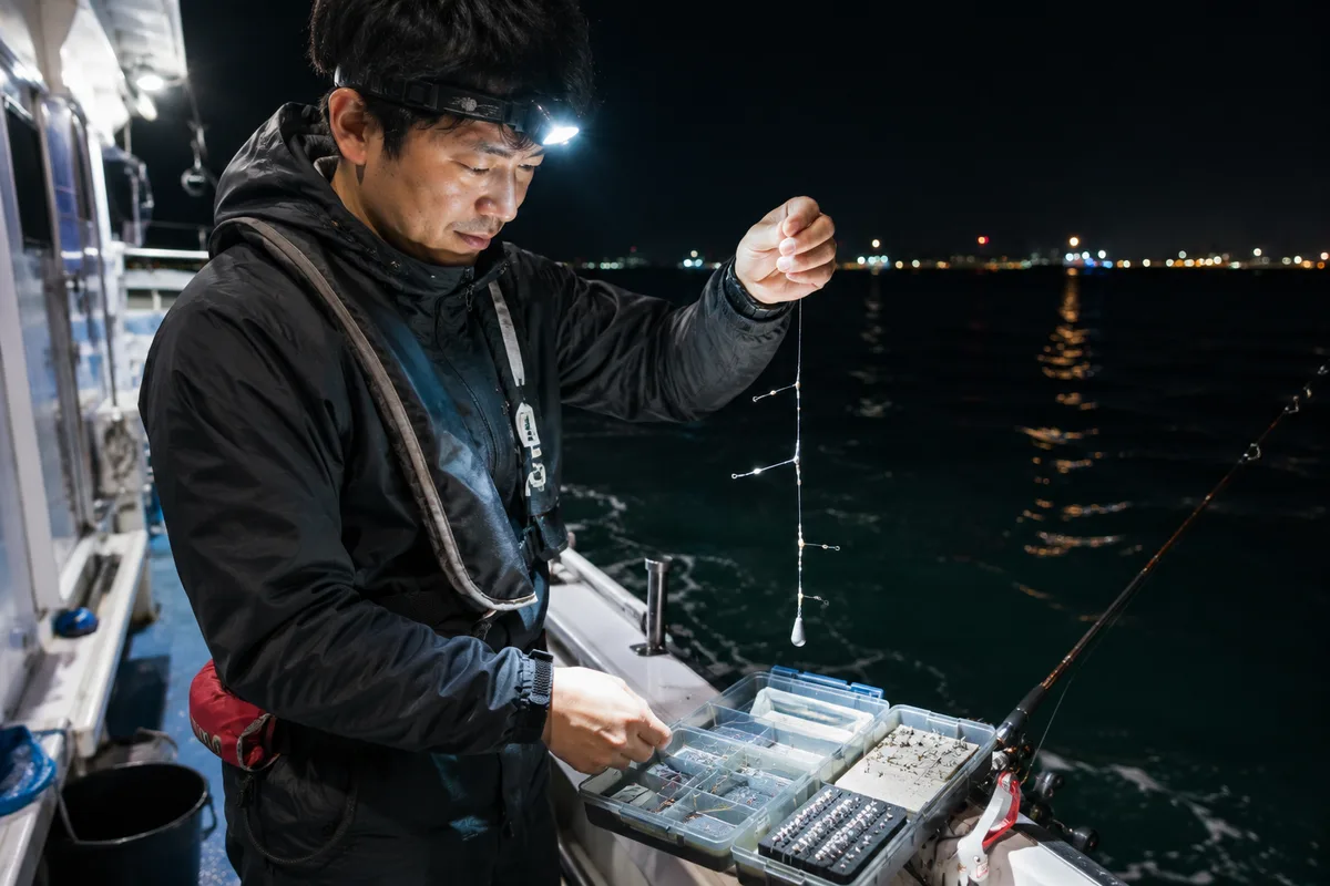 夜の遊漁船デッキでヘッドライトを装着し、仕掛けの準備をする釣り人。手元がライトで明るく照らされ、周囲は夜の海が広がる