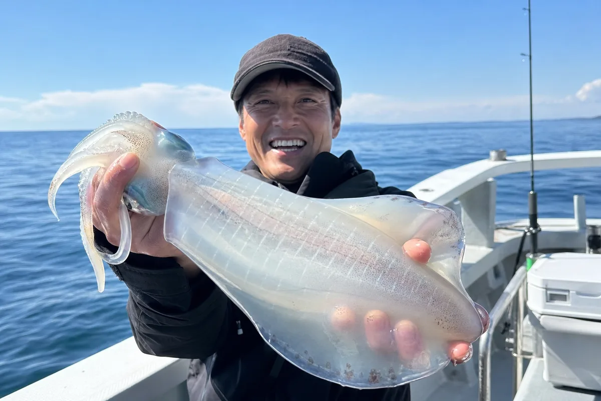 水面から抜き上げたアオリイカを掲げ、笑顔を見せる釣り人。エギング船のデッキを背景にした明るい写真