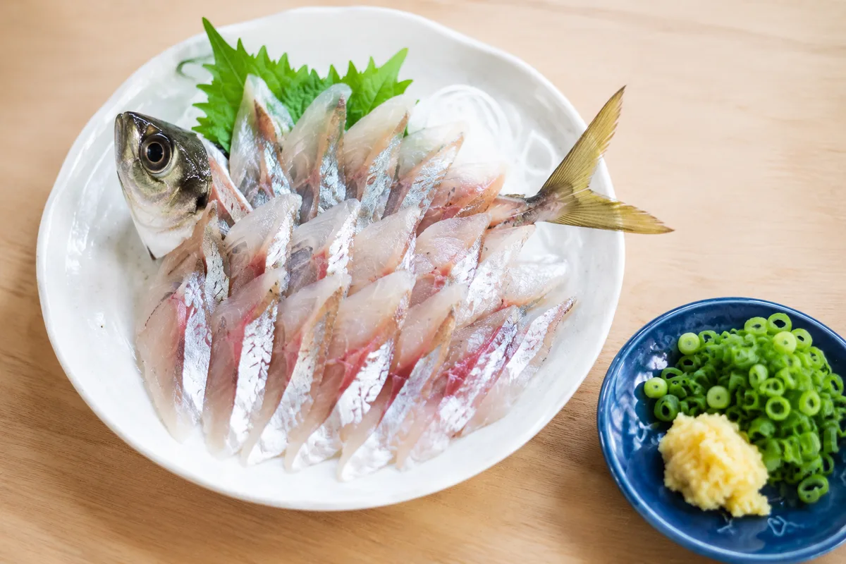 釣りたてのアジ（20〜25cm）を三枚にさばいた刺身と薬味が白い和皿に盛り付けられた様子。青い小皿にネギと生姜