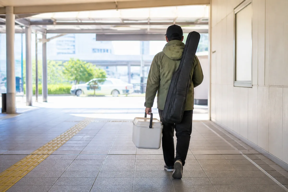 電車釣行の釣り人が、20L台のクーラーボックスとロッドケースを持って改札前を歩く場面