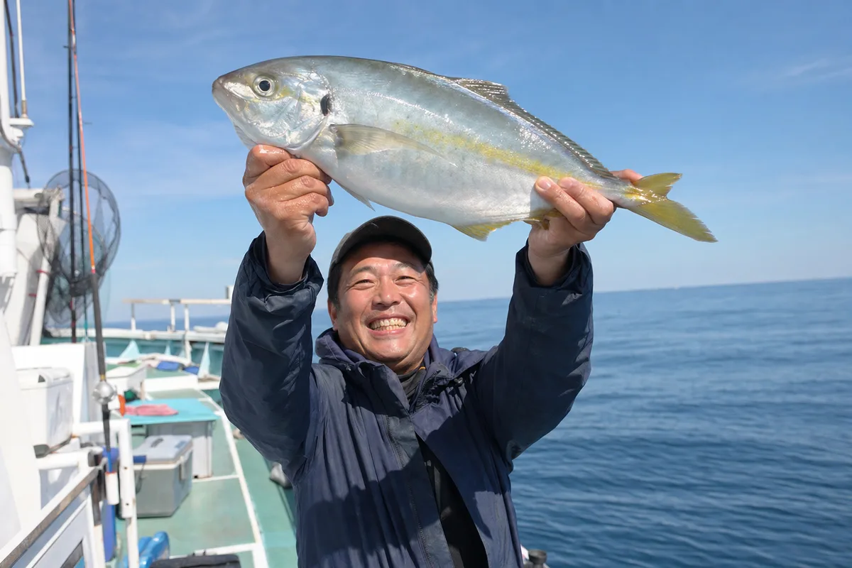 水面から抜き上げられた銀色に輝くシマアジを両手で持ち上げ、満面の笑みを見せる釣り人。房総の海を背景にした明るい写真